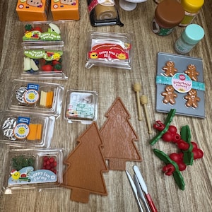 May include: A selection of miniature food items, including crackers, a roll cake, and deli items. There are also gingerbread biscuits, honey dippers, and decorative trees. The items are arranged on a wooden surface.
