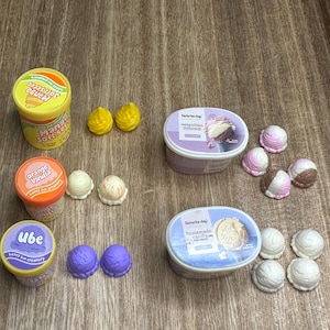 Pode incluir: Uma coleção de potes e bolas de sorvete de brinquedo. Os sabores incluem manga, laranja baunilha, ube, napolitano e baunilha caseira. Os potes são amarelos, laranja e roxos com bolas correspondentes.