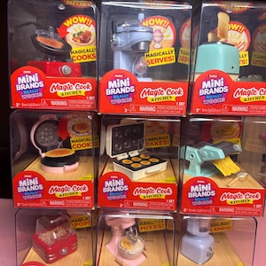 Op de afbeelding: Een verzameling ZURU Mini Brands Magic Cook Kitchen speelgoed in doorzichtige plastic verpakkingen. Het speelgoed omvat miniatuurversies van een koekenpan, mixer, broodrooster, wafelijzer en andere keukenapparaten. Elke doos heeft de tekst "REALLY WORKS".