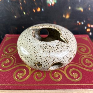 Peut inclure: Un cendrier en céramique mouchetée de forme circulaire avec plusieurs trous. Le cendrier est beige clair avec des mouchetures marron et une grande ouverture supérieure. Il repose sur un livre rouge avec des volutes décoratives dorées.