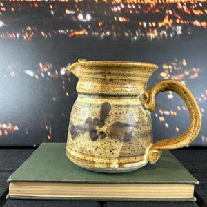 May include: A brown and tan ceramic jug with a speckled glaze and a brown handle. The jug is sitting on a green book.