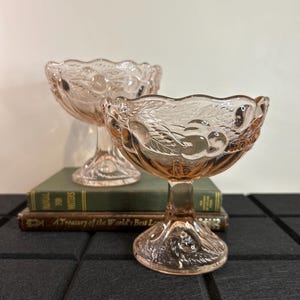 May include: Two pink glass pedestal bowls with a floral design. The bowls are on top of a stack of books.