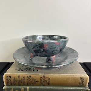 May include: A ceramic bowl with three small feet and a matching saucer, both with a mottled blue, pink, and grey glaze. The bowl sits atop a stack of two vintage books with gold and green covers.