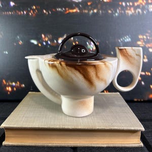 May include: A vintage ceramic teapot with an attached cup. The teapot is off-white with brown marbling and a dark brown lid and handle. The cup has a circular handle. The teapot is on a book.