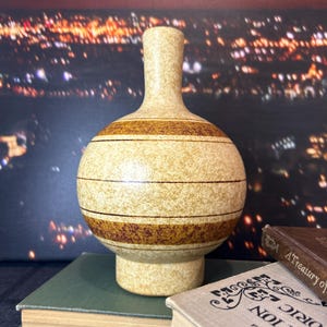 May include: A ceramic vase with a rounded body and a thin neck. The vase has a light brown glaze with brown stripes and speckles.