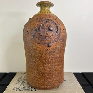 May include: A handcrafted, brown ceramic vase with a textured surface. The vase has a cylindrical body and a rounded top with a smaller, olive-green neck. The design includes a spiral pattern and small, raised details.