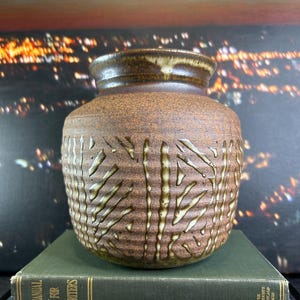 May include: A brown ceramic vase with a textured surface and a wide opening. The vase features vertical, textured lines in a lighter brown. The vase sits on two books with gold lettering. The books are green and blue.