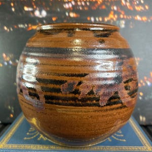 May include: A handcrafted, round ceramic vase with a brown and black striped design. The vase has a textured surface and a glossy finish. The vase is sitting on a blue book with gold lettering. The background is blurred with city lights.