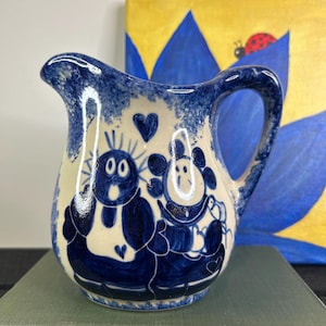 May include: A small ceramic jug with a blue and white design. The jug has a handle and spout, with a whimsical illustration of two cartoon characters. The background is a painting with blue and yellow colours.