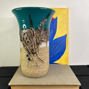 May include: A teal ceramic vase with a textured brown and white design of stylised branches and berries. The vase is sitting on a book.