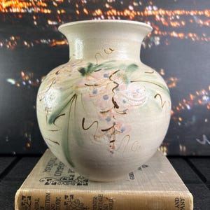 May include: A cream ceramic vase with a rounded body and flared rim. The vase features delicate floral patterns in pink, green, and brown. It is placed on a vintage book.