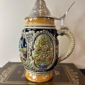 May include: A ceramic beer stein with a blue and white floral design, a silver lid, and a handle. The stein is decorated with a scene of a man and a woman in a garden.