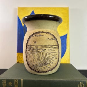 May include: A ceramic vase with a brown rim and a carved image of a lighthouse on a cliff overlooking the ocean. The vase is sitting on top of two books.
