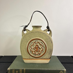 May include: A ceramic flask with a light green glaze, featuring a brown circular design on the front and a leather handle. The flask has two small handles and sits on a green book. The design is reminiscent of a vintage style.