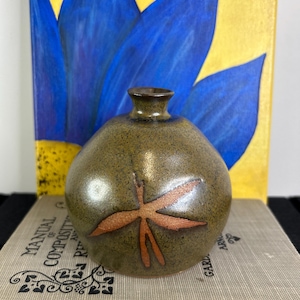 Peut inclure: Petit vase rond en céramique vert olive avec un bord marron et un motif de libellule orange stylisé. Le vase est présenté devant une peinture d'une fleur bleue et d'une coccinelle.