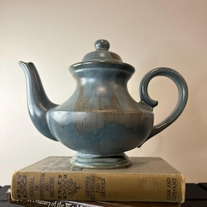 May include: A blue ceramic teapot with a brown glaze sits on top of a stack of three books. The top book has the title "A Treasury of the World's Best Loved Poems" and the author "Gardiner Kittredge and Arnold".