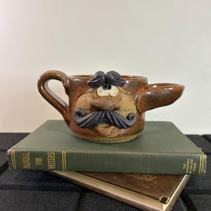 Peut inclure: Une tasse en céramique brune avec un visage, une moustache et des sourcils. La tasse est posée sur un livre vert dont le titre "Manuel pour les écrivains" est inscrit sur le dos.