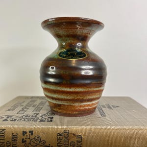Peut inclure: Petit vase artisanal en céramique avec un dessus évasé et un corps arrondi. Le vase présente une palette de couleurs marron, verte et crème avec des rayures horizontales. L'étiquette indique "Llangollen Pottery North Wales".