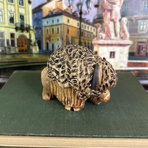 May include: A detailed figurine of a yak, with a textured, dark brown coat and a lighter brown body. The yak has a small, orange nose and black horns. The figurine is placed on a green book, with a blurred cityscape in the background.