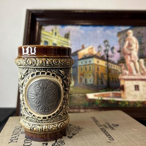 May include: A brown and cream ceramic beer stein with an ornate design and a silver-coloured emblem featuring a double-headed eagle. The stein is sitting on a book with the title "GARDENER AT THE BRIDGE" visible.