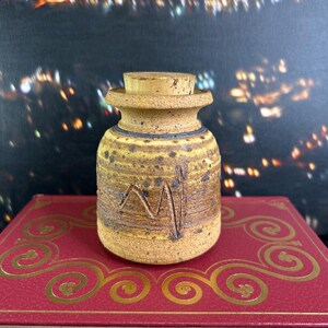 May include: A small, handcrafted ceramic jar with a cork stopper. The jar is tan with a textured surface, featuring dark brown speckles and etched lines. It sits on a red book with gold decorative swirls. The background is a blurred cityscape.