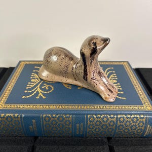 May include: A ceramic seal figurine in shades of brown and tan, with dark brown accents. The seal is posed in a resting position, with its head raised. The figurine is placed on a blue book with gold-coloured decorative elements.