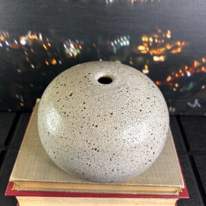 May include: A spherical ceramic vase with a speckled surface and a single hole at the top. The vase is light grey with dark brown speckles. It rests on a stack of books with red and gold detailing. The background is blurred.