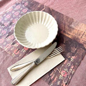 Pode incluir: Uma tigela de cerâmica branca com uma borda canelada está sobre um tapete de mesa estampado em marrom e rosa. Uma faca e um garfo de prata são colocados em uma toalha de mesa bege ao lado da tigela.