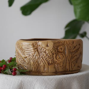 May include: A beige ceramic plant pot with a carved design of flowers and geometric shapes. The rectangular pot is displayed on a white cloth, with red berries and green leaves in the foreground.
