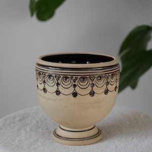 May include: A beige ceramic bowl with a dark brown interior and decorative black patterns. The bowl has a pedestal base and a detailed design around the rim. The bowl is likely used for decorative purposes or as a small planter.