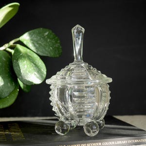 May include: A clear glass candy dish with a domed lid and a knob on top. The dish has a textured, faceted design and sits on three round feet.