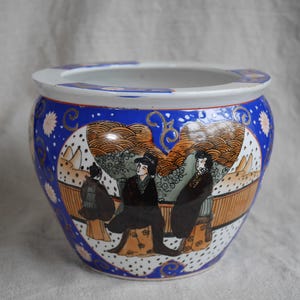 May include: A blue and white ceramic planter with a floral design and three figures in traditional Japanese clothing. The planter has a wide rim and a rounded body.