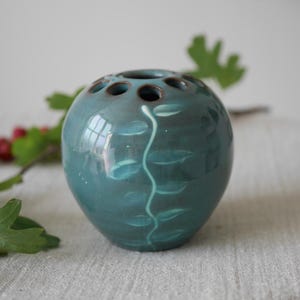 May include: Teal ceramic bud vase with a glossy finish. The vase has a round shape with several holes on top for flower stems. A white vine design with leaves decorates the front. A sprig of red berries and green leaves are in the background.