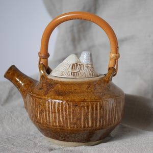 May include: A brown ceramic teapot with a white lid and a brown leather handle. The teapot has a textured surface and a unique design.