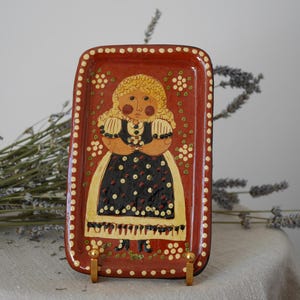 May include: A red ceramic tray with a white border and gold accents. The tray features a hand-painted illustration of a girl in a black and white polka dot dress holding a flower.