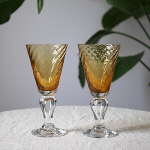 May include: A pair of amber-tinted stemmed glasses. The bowls have a textured, twisted design. The stems are clear with a decorative bulb shape. The glasses are on a white surface. Green leaves are in the background.