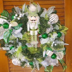 May include: A green and white Christmas wreath with a nutcracker figurine in the center. The wreath is decorated with green and white ribbon, green and white mesh, and white berries.