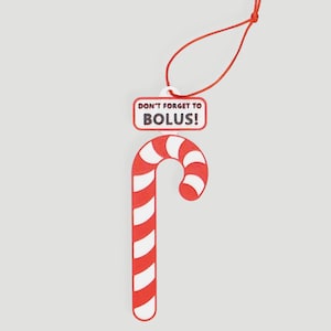 Peut inclure: Ornement de canne en bonbon rayée rouge et blanche, suspendu par une ficelle rouge. Une étiquette rectangulaire au-dessus de la canne indique "DON'T FORGET TO BOLUS!" en rouge. Fond gris clair.
