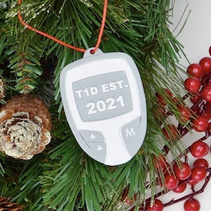 Peut inclure: Ornement de moniteur de diabète gris et blanc avec le texte "T1D EST. 2021" accroché à une couronne de Noël. La couronne est décorée d'aiguilles de pin, de pommes de pin et de baies rouges.