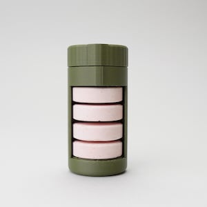 Puede incluir: Un pastillero cilíndrico verde oliva con tapa de rosca. El recipiente contiene cuatro pastillas rosas y circulares. El diseño es simple y moderno, con un enfoque en la funcionalidad y una estética limpia.