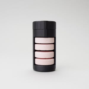 Puede incluir: Un recipiente de plástico negro con cinco tabletas rosadas apiladas en su interior. El recipiente tiene una tapa de rosca.