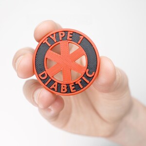 May include: A round, red and black 3D-printed emblem with the words "TYPE 1 DIABETIC" in white lettering. The emblem features a red star-shaped design in the center. The emblem is held in a hand.
