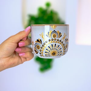 Pode incluir: Caneca de cerâmica branca com um design de mandala dourado e preto. A caneca está sendo segurada por uma mão com esmalte de unha rosa.
