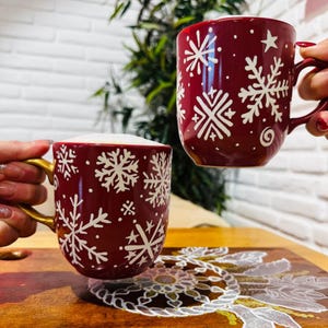 Op de afbeelding: Twee rode mokken met gouden handvatten en witte sneeuwvlokontwerpen. De mokken zijn gevuld met een warme drank.