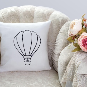 May include: White throw pillow with a black outline of a hot air balloon. The pillow is sitting on a cream-colored armchair with a floral arrangement in the background.