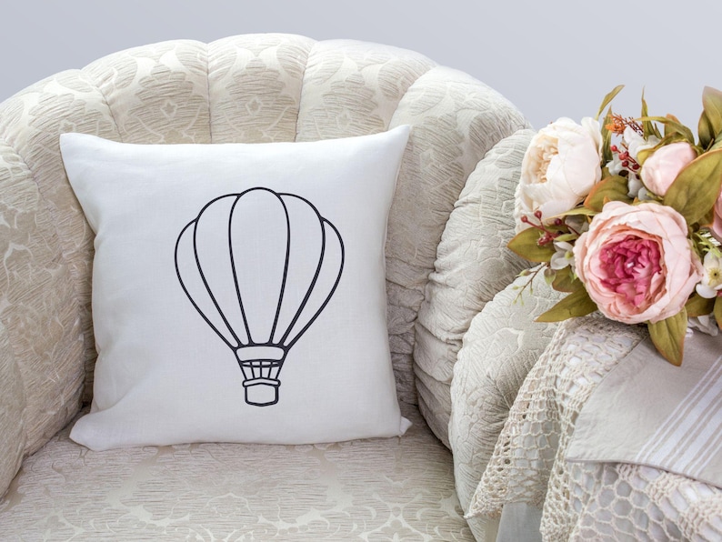 May include: White throw pillow with a black outline of a hot air balloon. The pillow is sitting on a cream-colored armchair with a floral arrangement in the background.