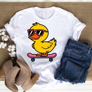 May include: A white t-shirt with a cartoon image of a yellow duck wearing sunglasses and riding a pink skateboard.