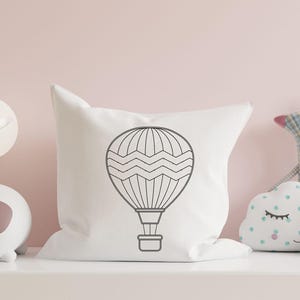 May include: A white pillow with a black outline of a hot air balloon with a wavy pattern. The pillow is on a light pink surface.