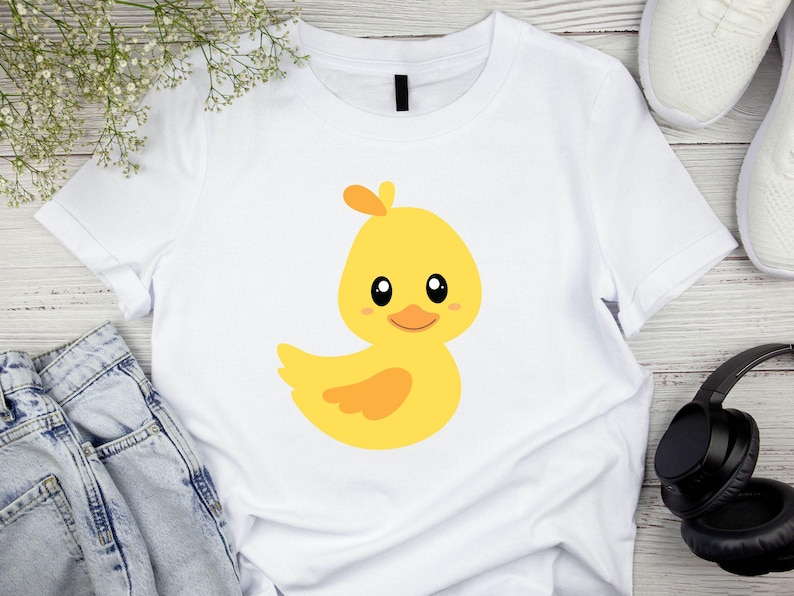 May include: A white t-shirt with a yellow cartoon duckling with orange accents. The duckling has big eyes and a small beak.