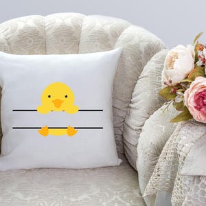 May include: White pillow with a yellow duckling peeking over a black line. The duckling has a black beak and orange feet. The pillow is on a cream-colored couch.